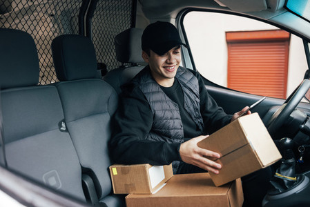 Smiling courier sitting on a drivers seat checking delivery information on mobile phone while holding box