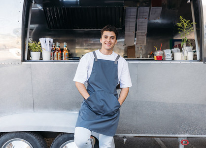 Portrait of a confident waiter in apron leaning on food truck and looking at camera