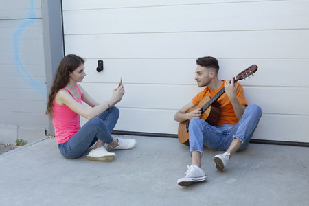 Young woman photographing boyfriend playing guitar in driveway