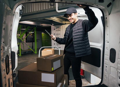 Smiling courier standing at the trunk of a van checks cardboard boxes on a cart using a digital tablet while standing in a warehouse
