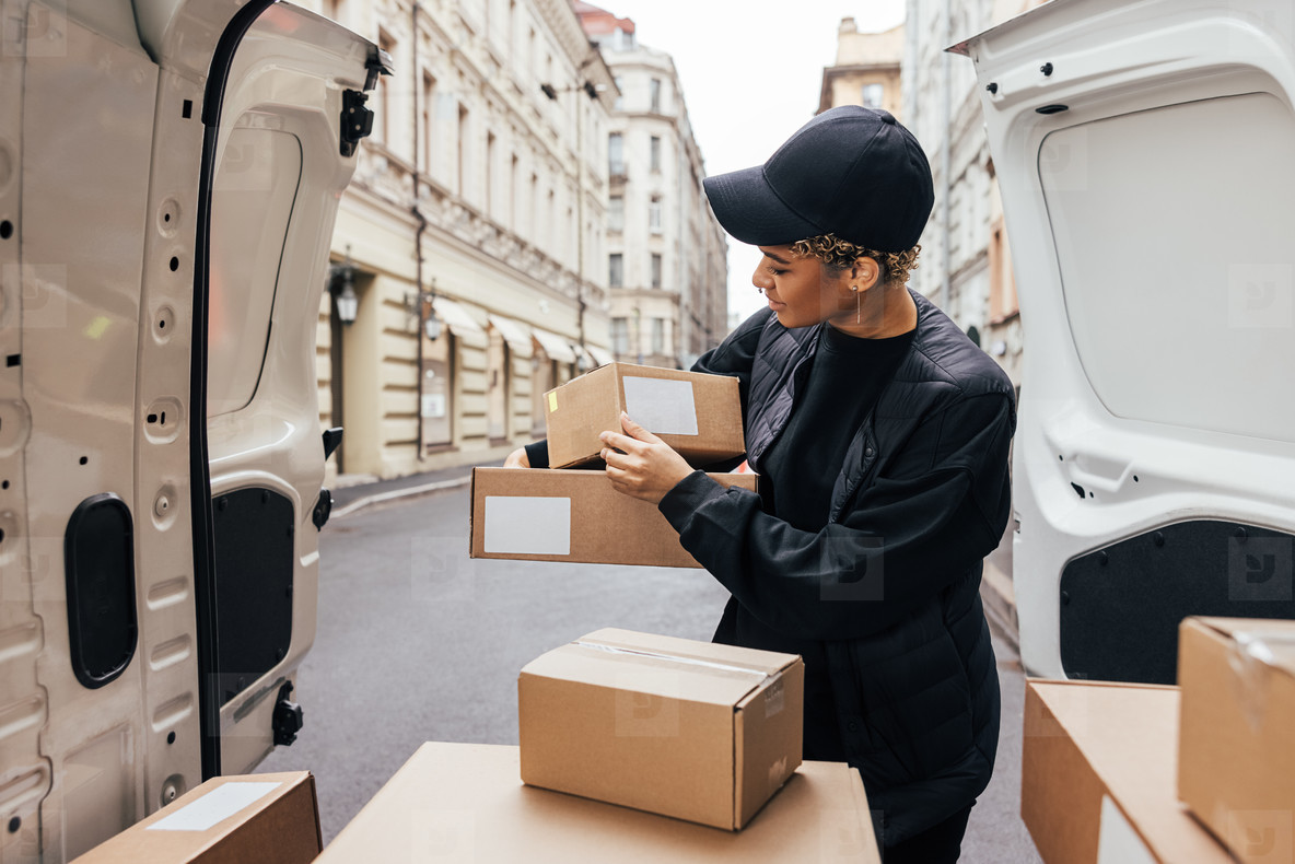 Woman in uniform holding packages while standing at car trunk  Courier preparing parcels for delivery