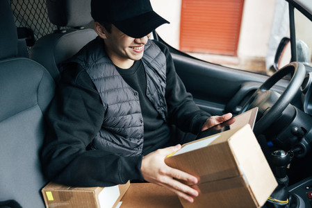 Smiling male courier in uniform sitting on drivers seat holding a package and looking on a digital tablet
