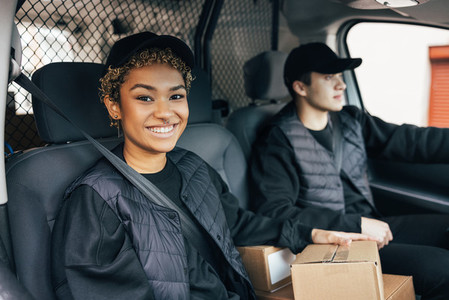 Smiling woman in uniform sitting in delivery van and looking at camera while her coworker driving