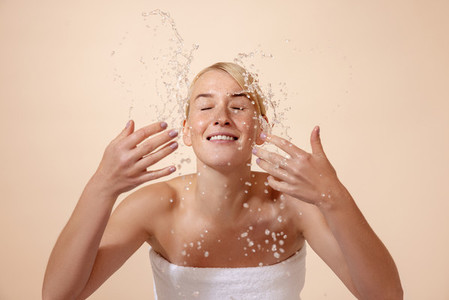 Cheerful blonde woman in white towel splashing water on her face