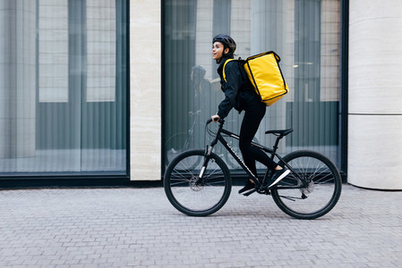 Side view of a young woman riding her bike in the city  Courier with a thermal backpack in her way to deliver food to a customer