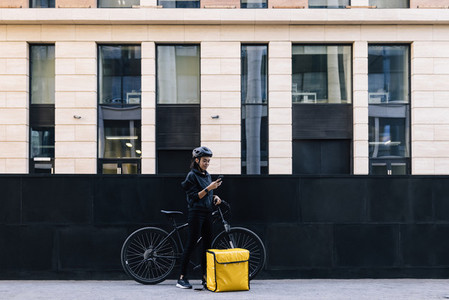 Side view of delivery woman holding a mobile phone  wearing a cycling helmet  Young female standing in the city with bicycle and thermal backpack