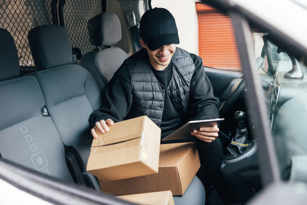 Smiling male courier in uniform and cap checking information on cardboard box while sitting in a car using a digital tablet