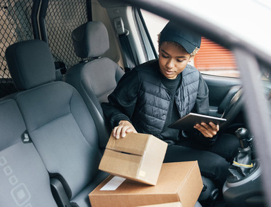 Woman courier checking information on cardboard box while sitting in a car using a digital tablet