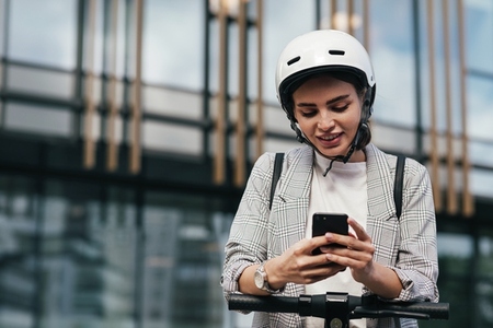Woman in formal wear typing on smartphone while leaning on the handlebar of electric scooter while standing at an office building
