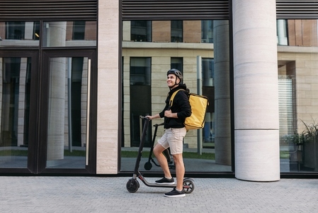 Smiling messneger man standing outdoors with electric push scooter  Delivery man with termal backpack in the city