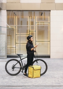 Side view female messenger standing outdoors with bicycle and thermal backpack holding a smartphone looking for a customer address
