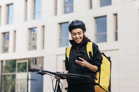 Female courier in cycling helmet with bicycle in the city  Smiling woman with thermal backpack looking at smartphone