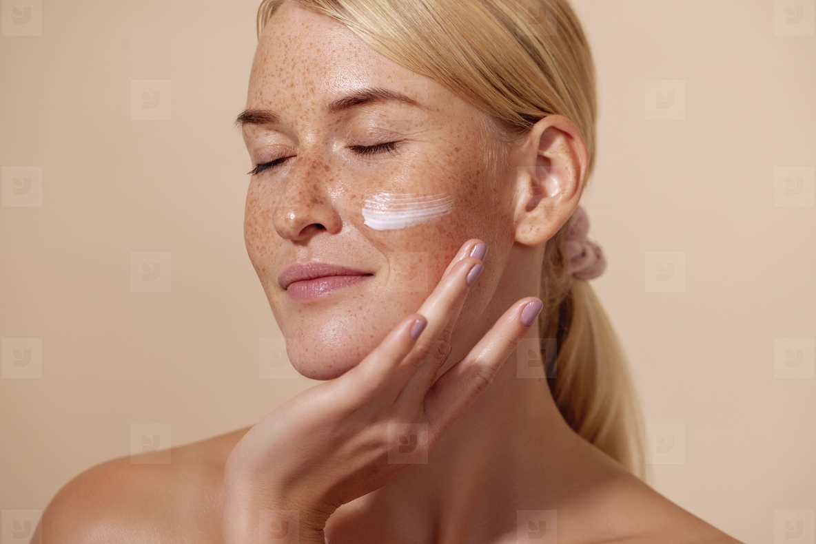 Close up of beautiful young female with cream on a face Smiling woman with closed eyes touching cheek with fingers