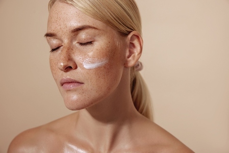Close up of a calm and beaautiful blond woman with facial cream on a cheek against pastel background