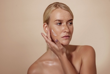 Confident woman with freckles applying anti aging cream on a cheek in studio while looking away