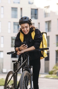 Smiling woman messenger with bicycle and thermal backpack holding a smartphone in the city
