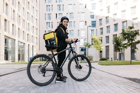 Smiling courier girl with bicycle  wearing a delivery thermal backpack looking at camera