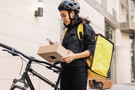 Side view of female messenger wearing cycling helmet and backpack looking on mobile phone while holding a parcel