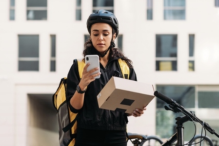 Delivery woman in cycling helmet holding a cardboard box checking information on mobile phone