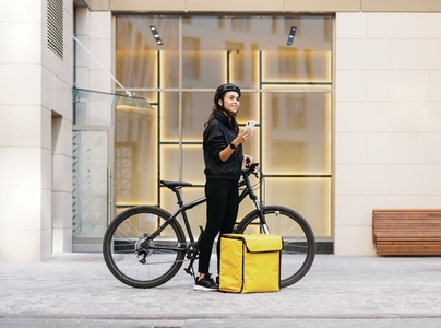Side view of woman in uniform with bicycle and food delivery backpack holding a mobile phone while standing at apartment building