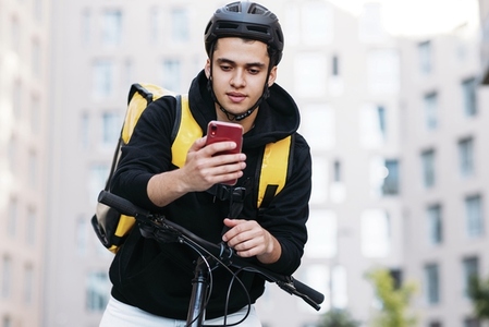 Male courier leaning on the handlebar of a bicycle wearing food delivery backpack and cycling helmet looking at mobile phone