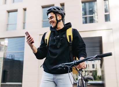 Smiling delivery man walking with bicycle looking on mobile phone in the city