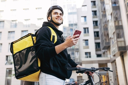 Portrait of delivery man holding a mobile phone  wearing thermal backpack standing against apartment building