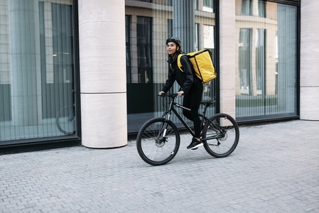 Smiling courier girl riding a bicycle wearing delivery thermal backpack looking for a customer address