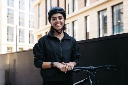 Young cheerful woman in cycling helmet standing in the city with a bike and looking at camera