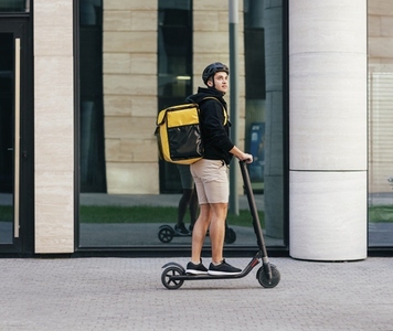 Side view of male courier riding on electric push scooter with food delivery backpack