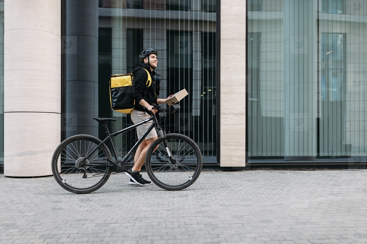 Side view of a male courier walking with a bicycle holding a package  Delivery man with thermal backpack at an office building