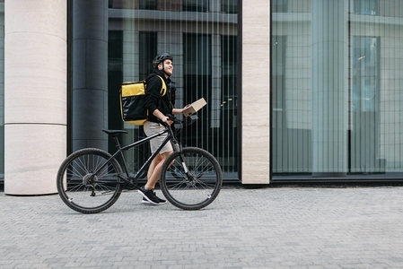 Side view of a male courier walking with a bicycle holding a package  Delivery man with thermal backpack at an office building