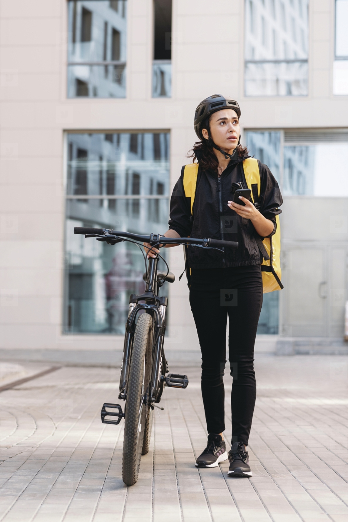 Woman courier walking on a city street with bicycle  holding mobile phone looking away