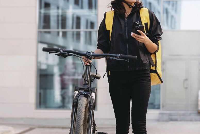 Woman courier walking on a city street with bicycle, holding mobile phone looking away