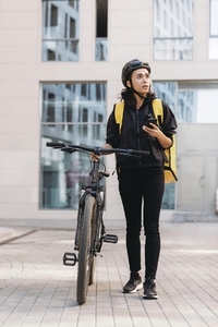 Woman courier walking on a city street with bicycle  holding mobile phone looking away