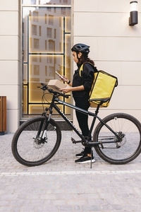 Side view of woman courier with backpack and bicycle standing at apartment building holding a cardboard box