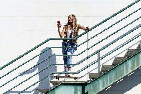 Black woman with coloured braids  looking at her smartphone with her feet resting on a skateboard