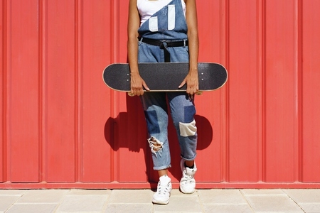 Unrecognizable woman holding a skateboard with her hands on a red urban wall