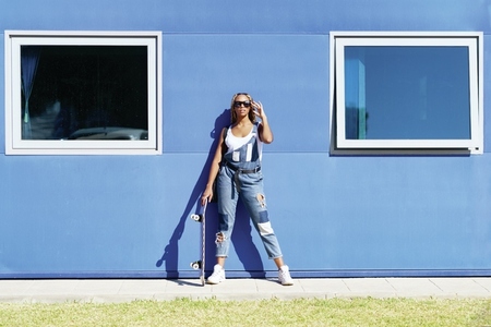 Black woman dressed casual  with modern sunglasses and a skateboard on blue wall background