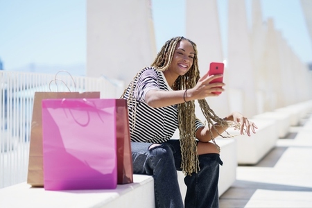 Black woman taking a selfie with her smartphone sitting on a bench in the street