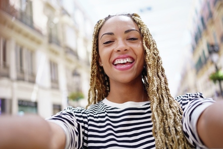 Black woman with afro braids taking a smiling selfie in an urban street with a smartphone