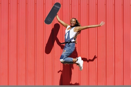 Black woman dressed casual wtih a skateboard jumping with happiness on red urban wall background