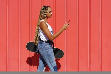 Black woman with coloured braids  consulting her smartphone with her feet resting on a skateboard