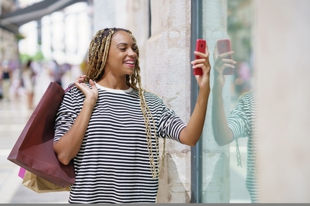 Young black woman photographing a store window in a shopping street with her smartphone