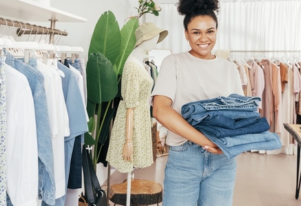 Smiling female fashion store worker holding a pile of jeans and looking at camera
