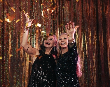 Two smiling senior women in black dresses throwing colorful confetti while standing against a golden backdrop in studio