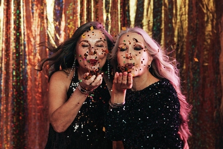 Two senior women in black dresses blowing confetti off their hands against a golden background