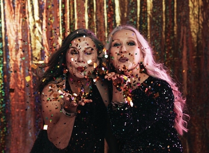Two fashionable senior women in black dresses blowing confetti off their hands in studio
