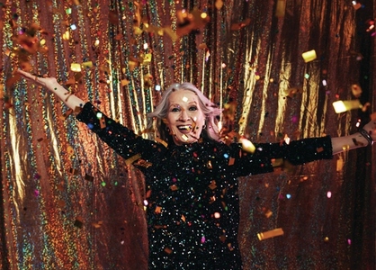 Smiling senior woman in black dress having fun under colorful confetti against a golden backdrop