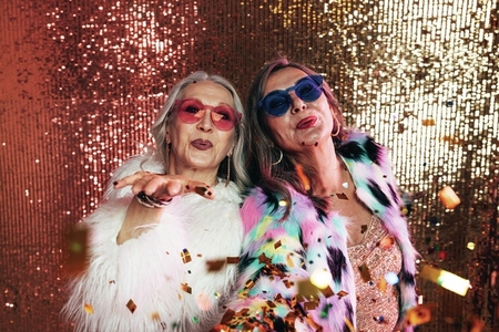 Two senior women in eyeglasses wearing fur coats blowing confetti off their hands against glitter background